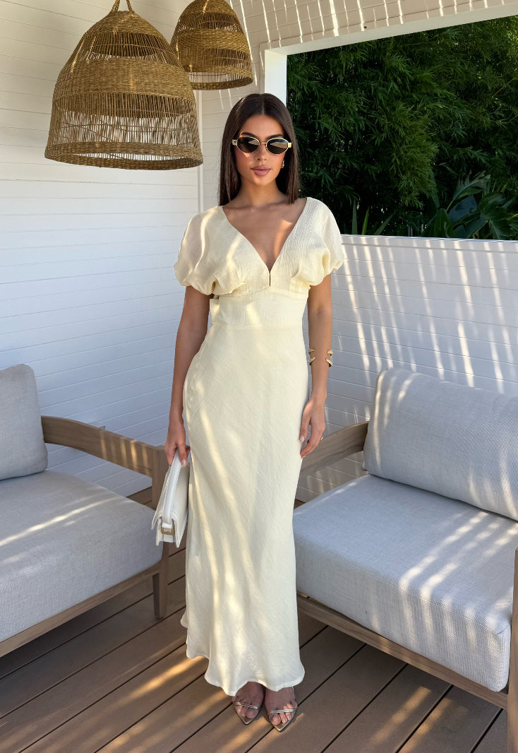 Woman in a lemon dress standing on a sunlit patio with wicker lanterns and outdoor furniture.