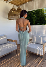 Woman in a light sage dress  thats backless standing in an outdoor setting with white furniture and greenery.